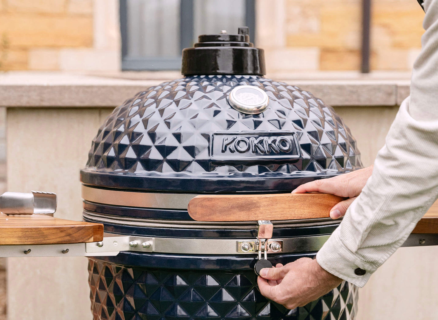 Hand operating the safety latch of a black Kokko kamado, featuring a textured ceramic lid, wooden handle, and built-in thermometer, in outdoor use.