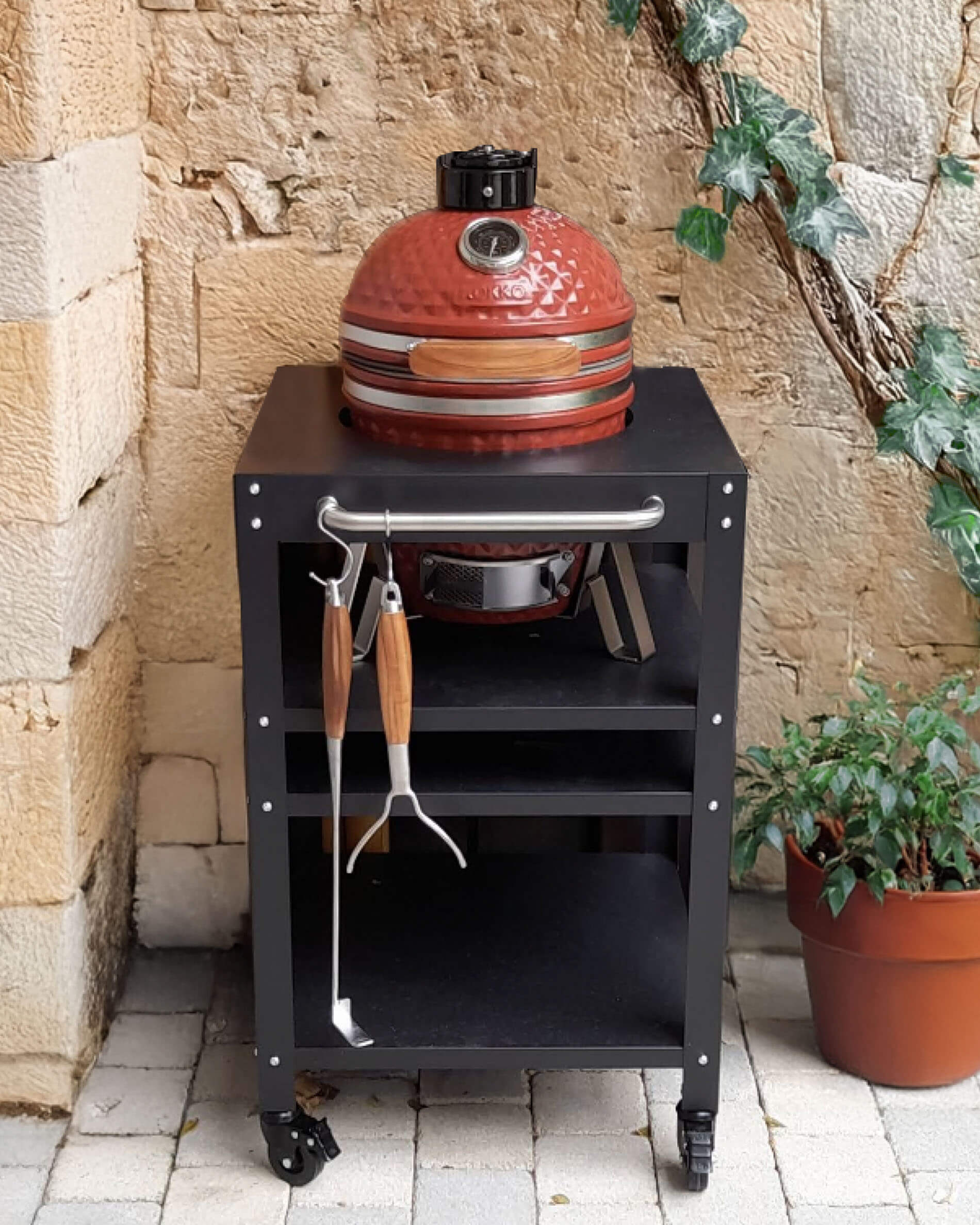 Red Kokko ceramic kamado set on a black wheeled cart, featuring an upper shelf, open storage space, and utensils hanging at the front, displayed in a stone courtyard.