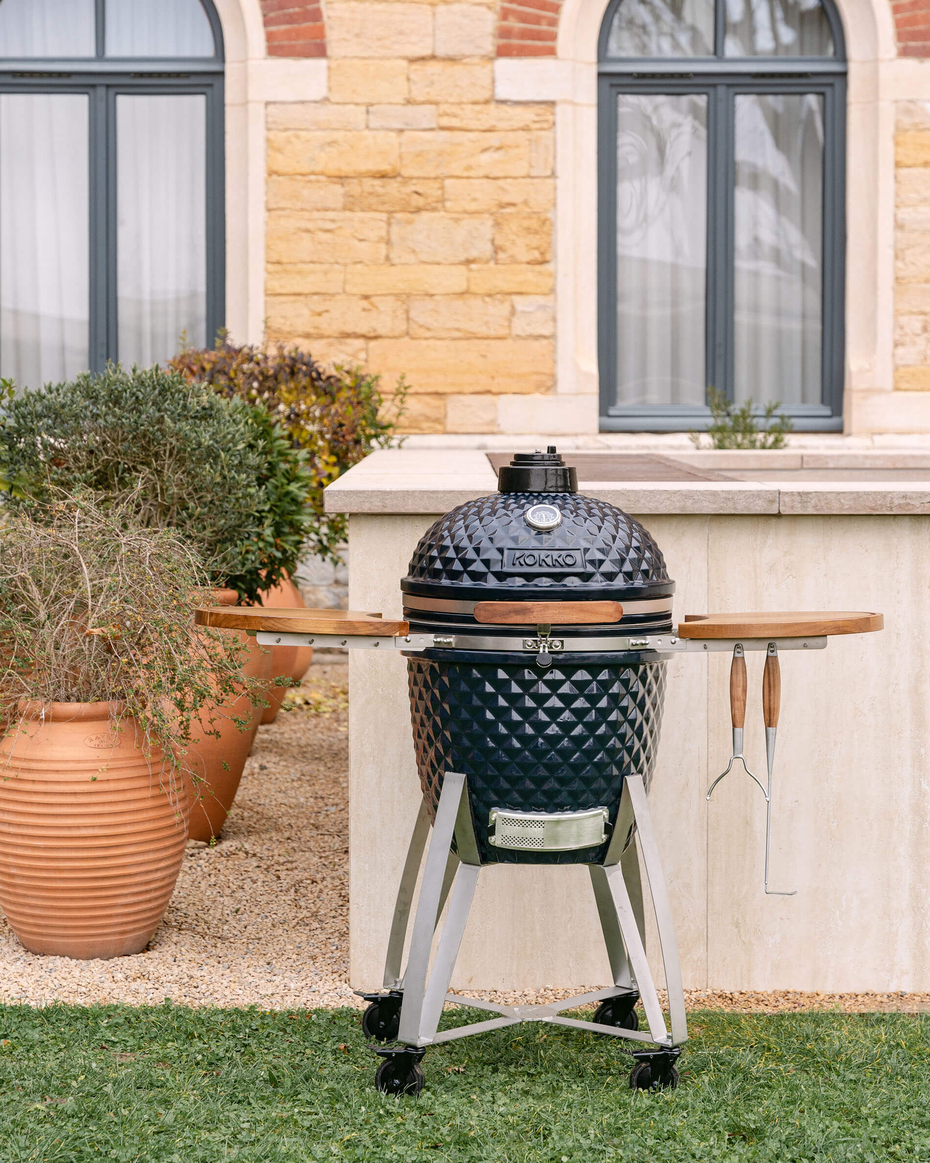 Overview of the Kokko kamado on a mobile cart with folding side shelves and hanging utensils, set up outdoors.