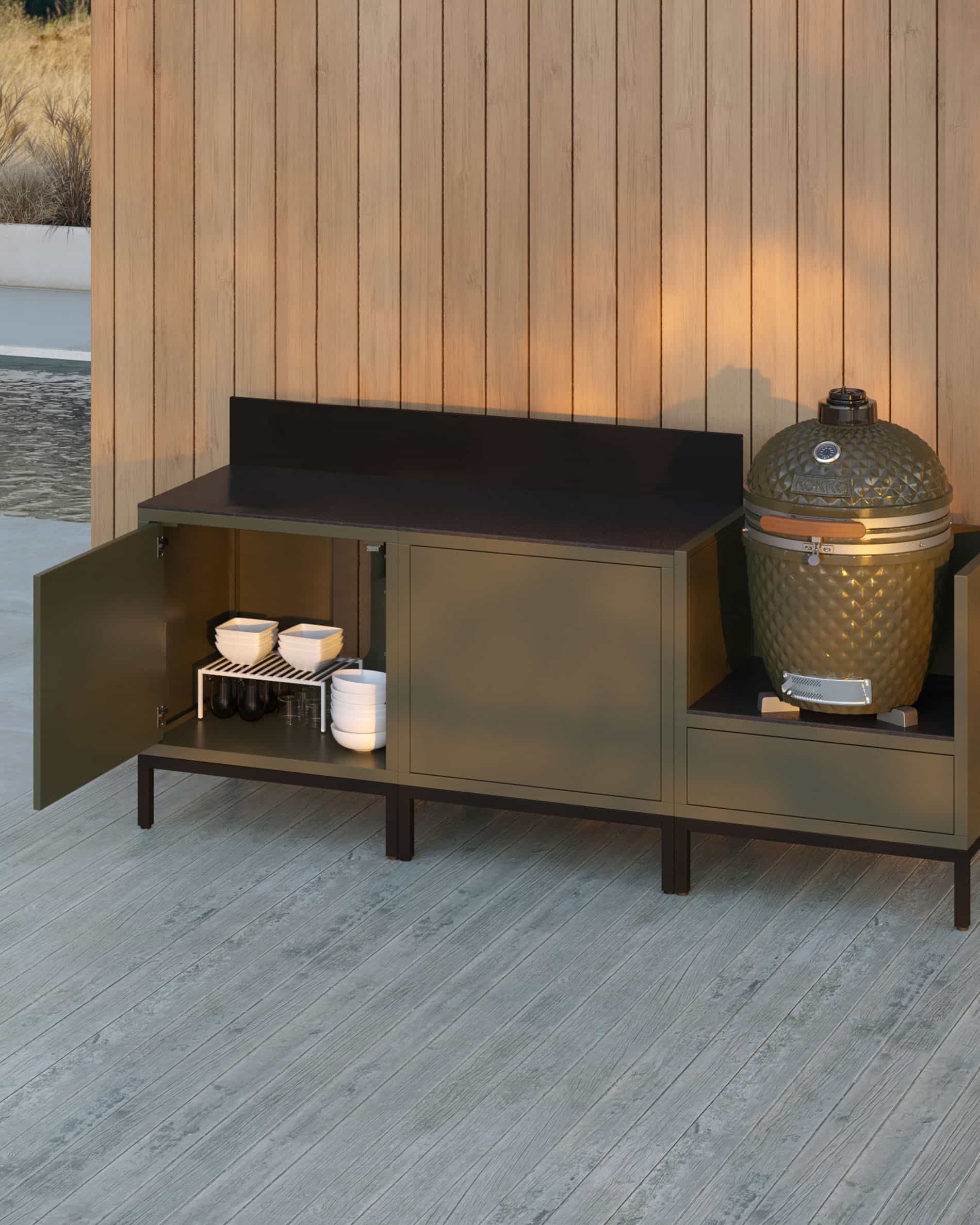 Cuisine pilotis 3 modules pour kamado, meuble coloris vert olive avec plan de travail sombre et crédence assortie, porte de rangement ouverte révélant vaisselle stockée, kamado installé dans le module latéral droit, structure sur pieds noirs.