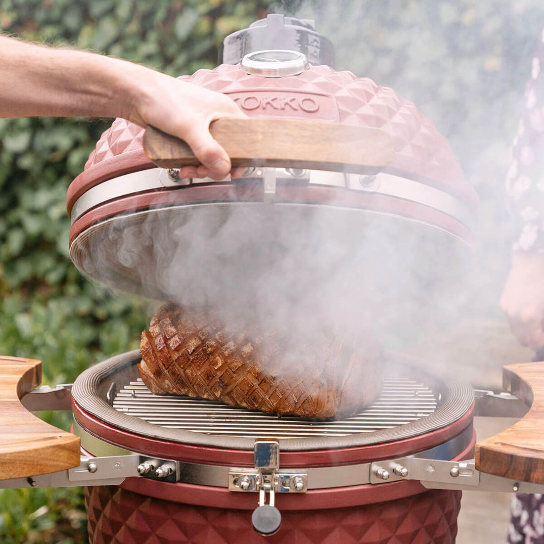A hand lifts the lid of a Kokko Oxide Red kamado, revealing a grilled piece of meat on the stainless steel grate. Thick smoke rises from the chamber, highlighting the wood-fired cooking, the peripheral seal, and the wooden side shelves.