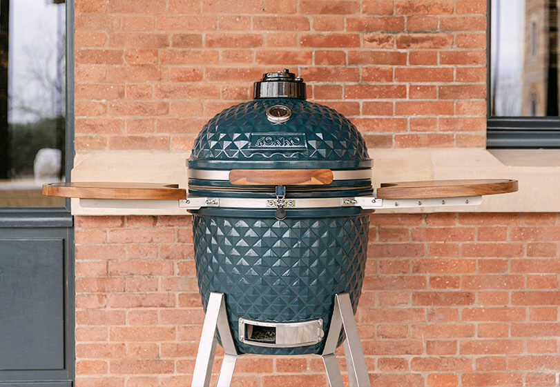 Ocean blue Kokko kamado set on a stand with wooden side shelves, shown from the front outdoors against a brick wall.