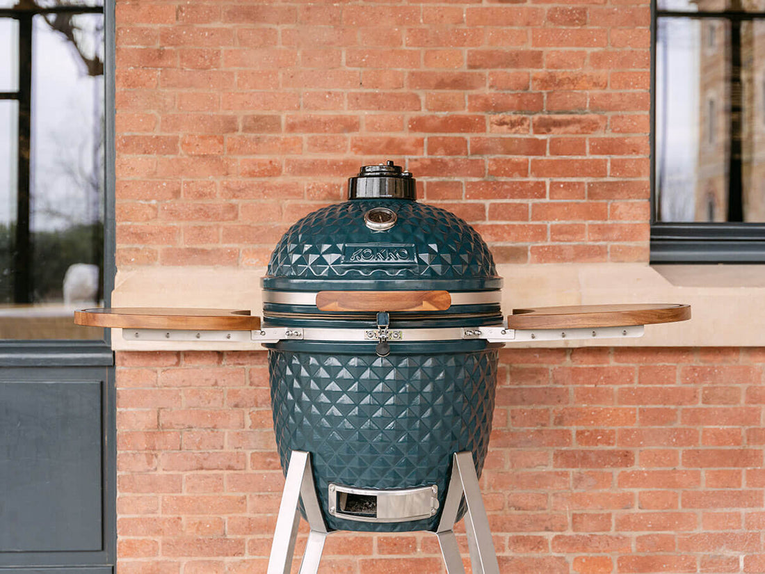 Ocean green Kokko kamado set up outdoors in front of a brick wall. The barbecue is closed, featuring a textured ceramic lid, an integrated thermometer on top, a wooden handle, a stainless steel safety latch, and two acacia wood side shelves. The whole unit stands on a metal frame with a visible lower ventilation tray.