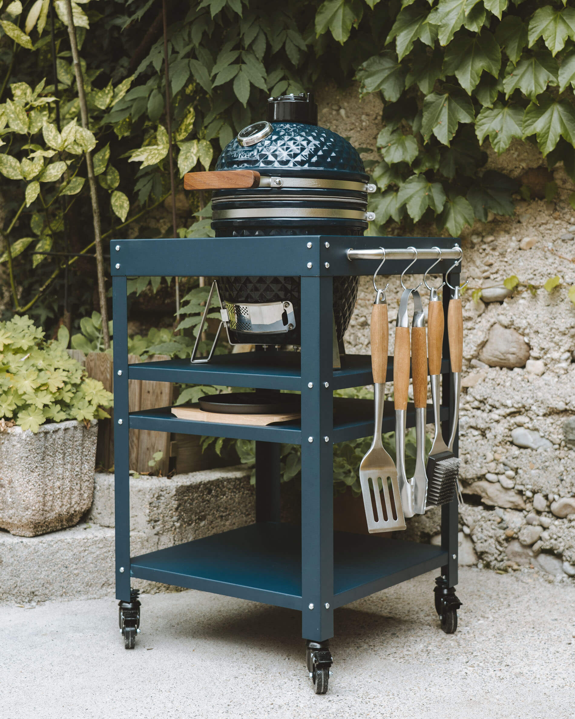 Kokko kamado in ocean blue set on a matching metal cart, featuring storage shelves, ash tray, and utensils hanging on the side.