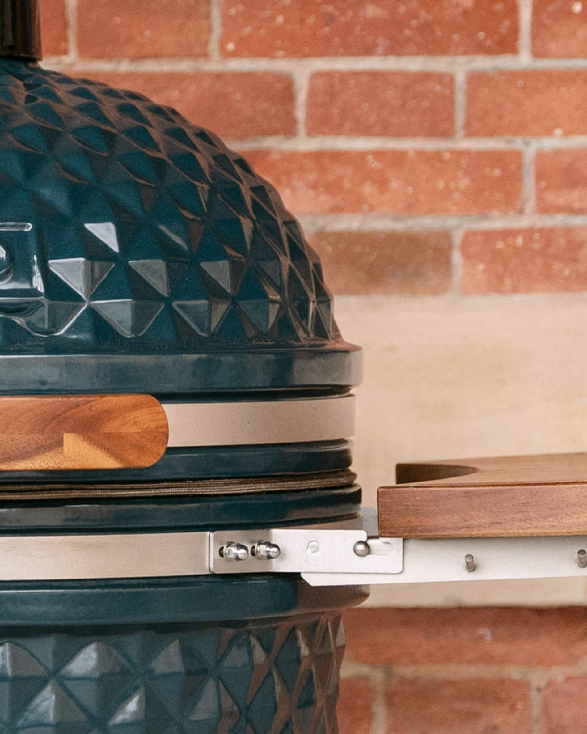 Close-up of the metal fittings connecting a wooden side shelf to the kamado body, with visible screws and gasket.