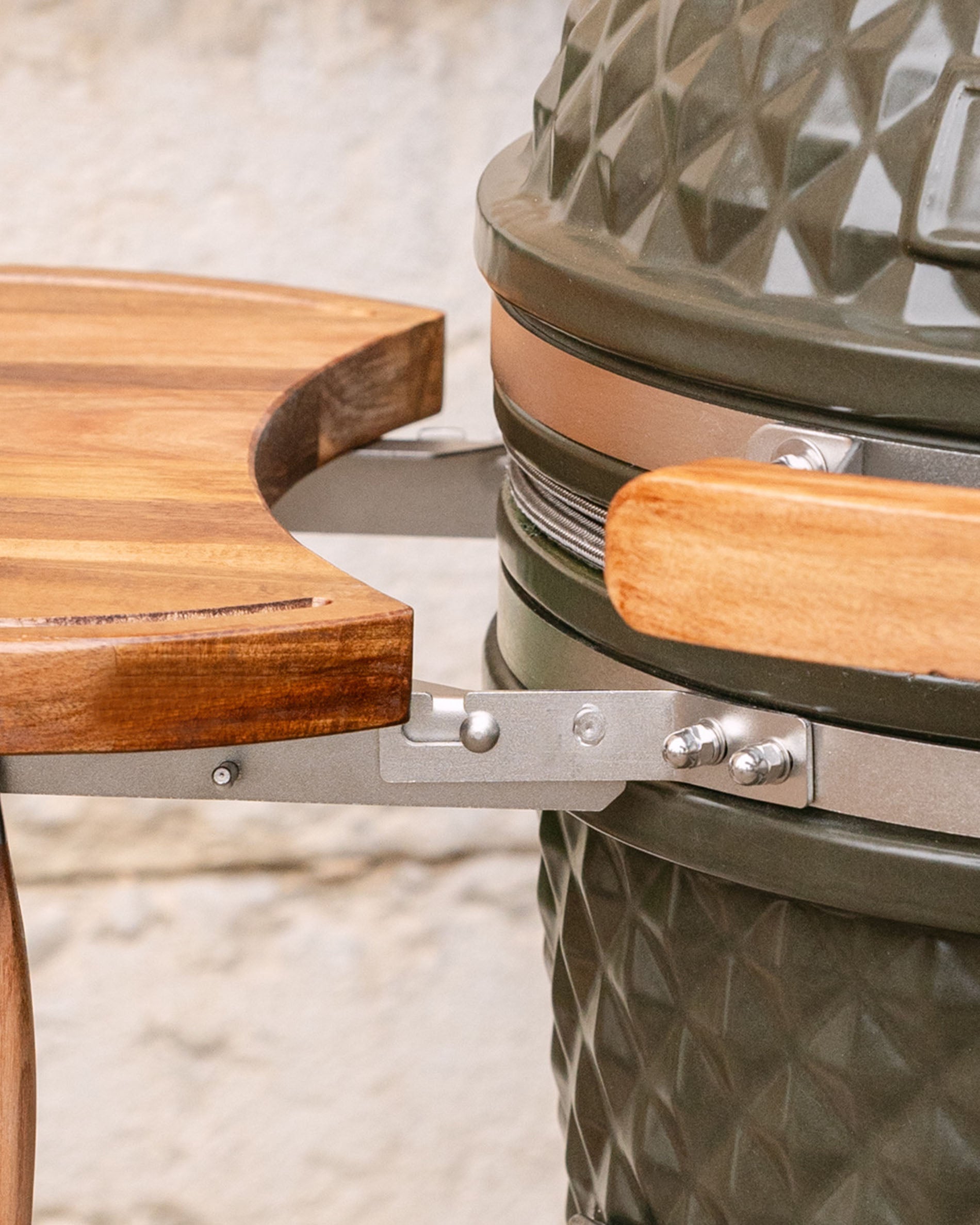 Close-up of the metal fittings connecting the wooden side shelf to the kamado, with visible wooden handle and stainless steel band.