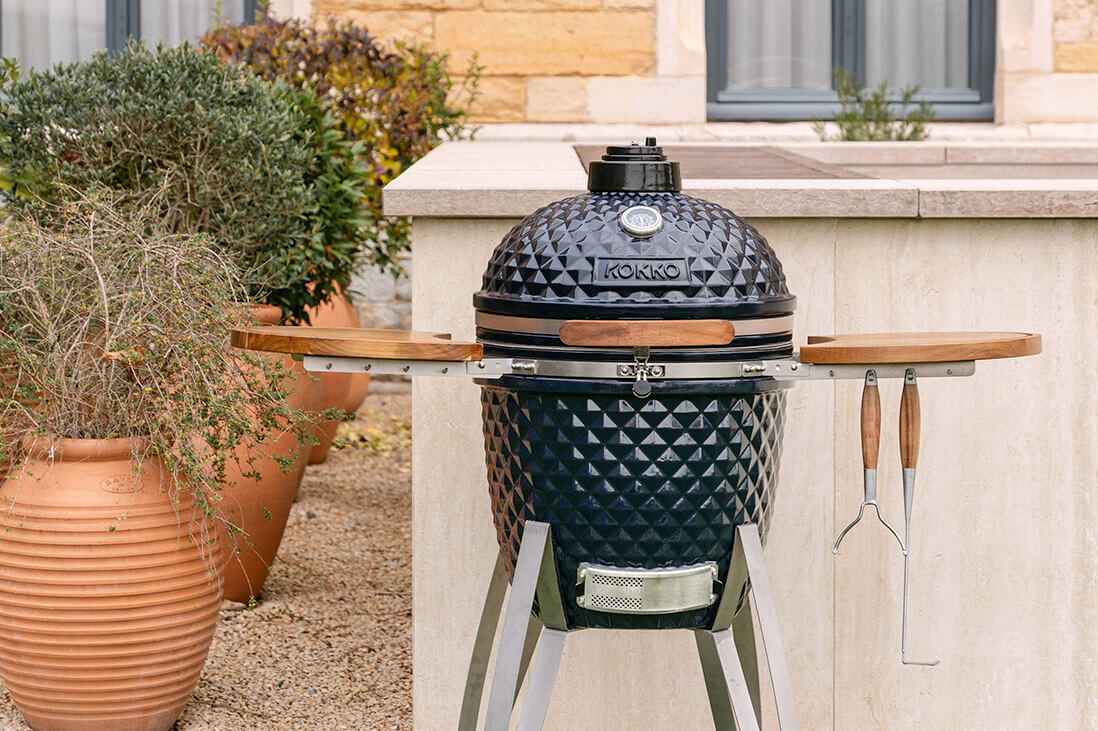 Kamado Kokko set up outdoors, featuring textured black ceramic, wooden side shelves, hanging utensils, and a metal frame, integrated into a garden kitchen space.