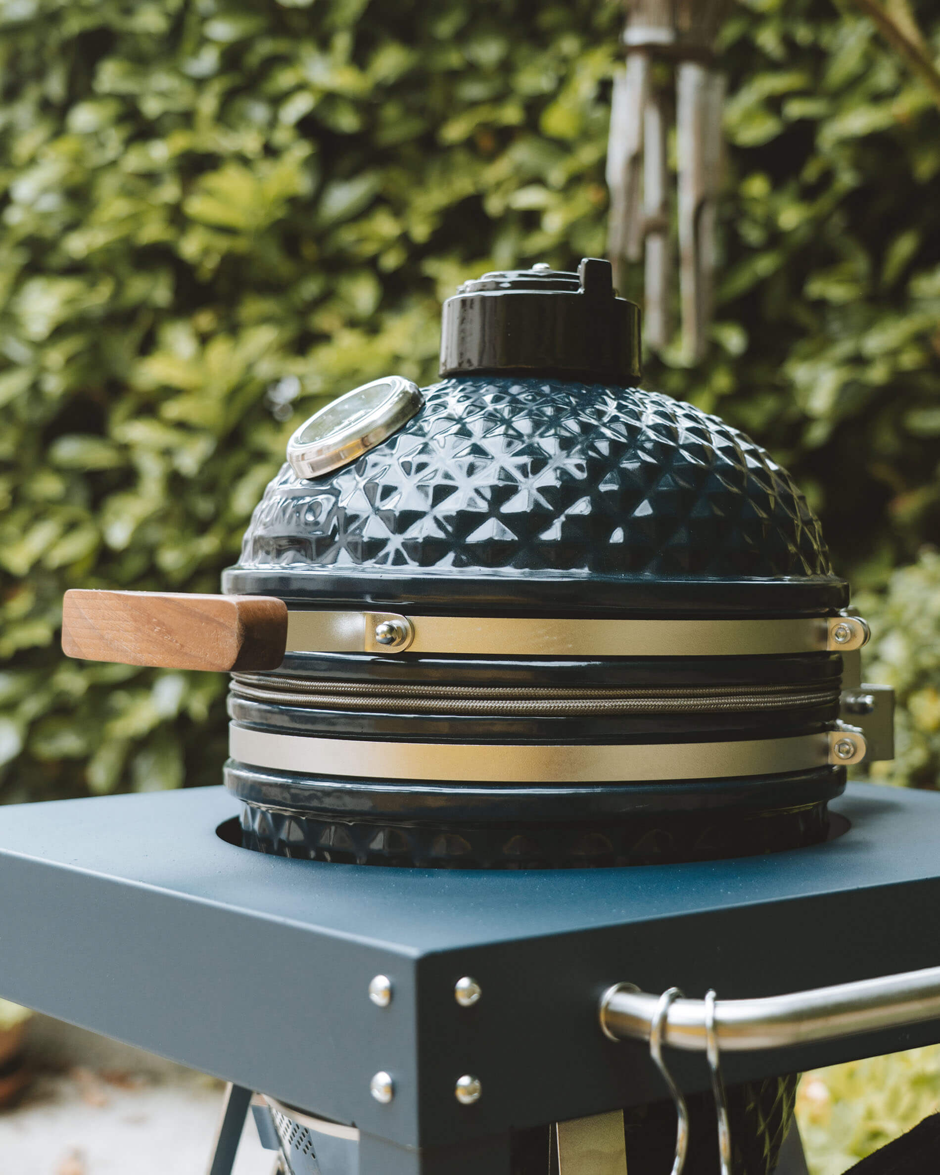 Vue latérale d’un kamado Kokko coloris océan installé sur chariot, mettant en valeur le couvercle en céramique, la poignée en bois et le thermomètre intégré.
