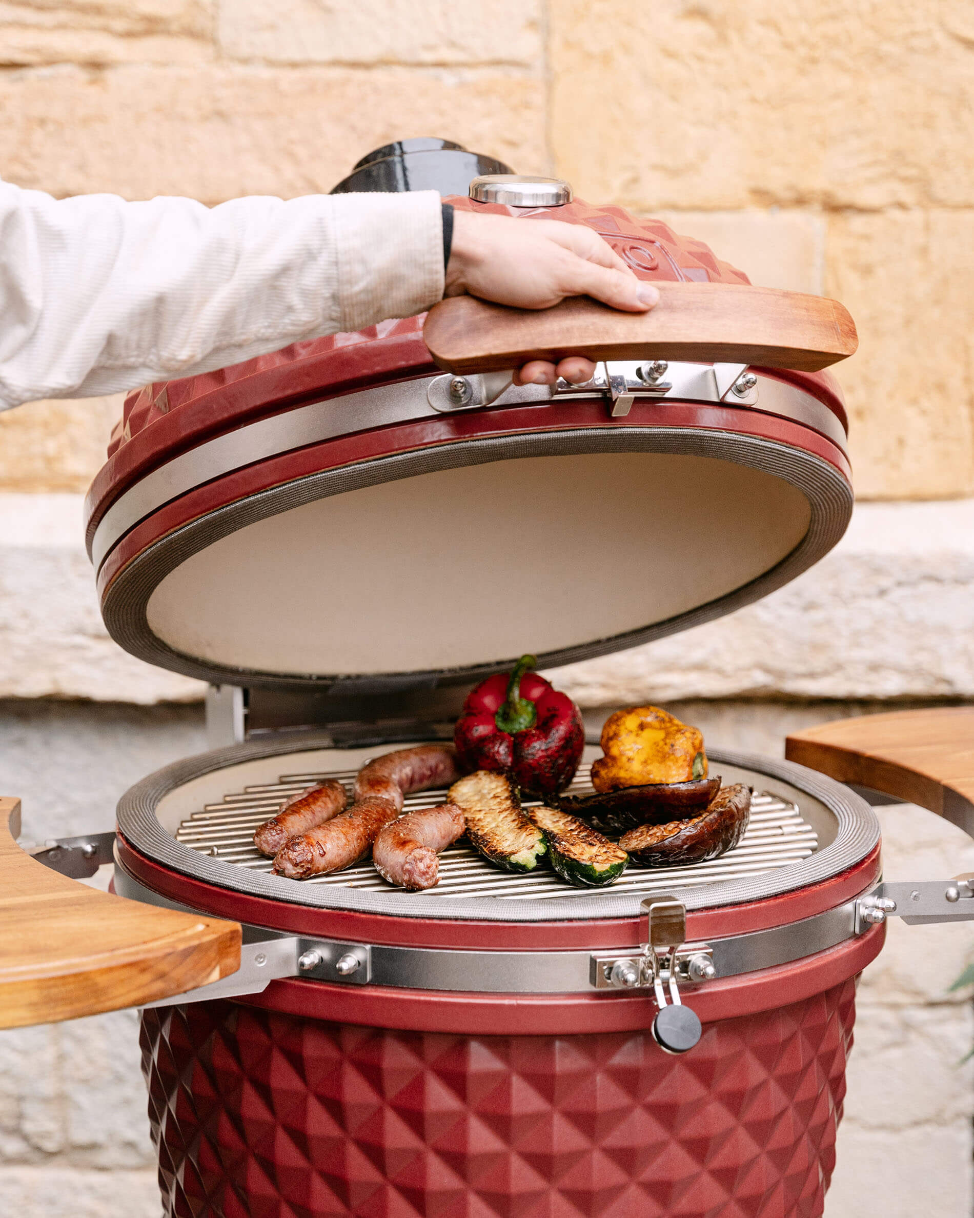 Red oxide ceramic Kokko 22-inch kamado open, grilling sausages and vegetables on the grate.