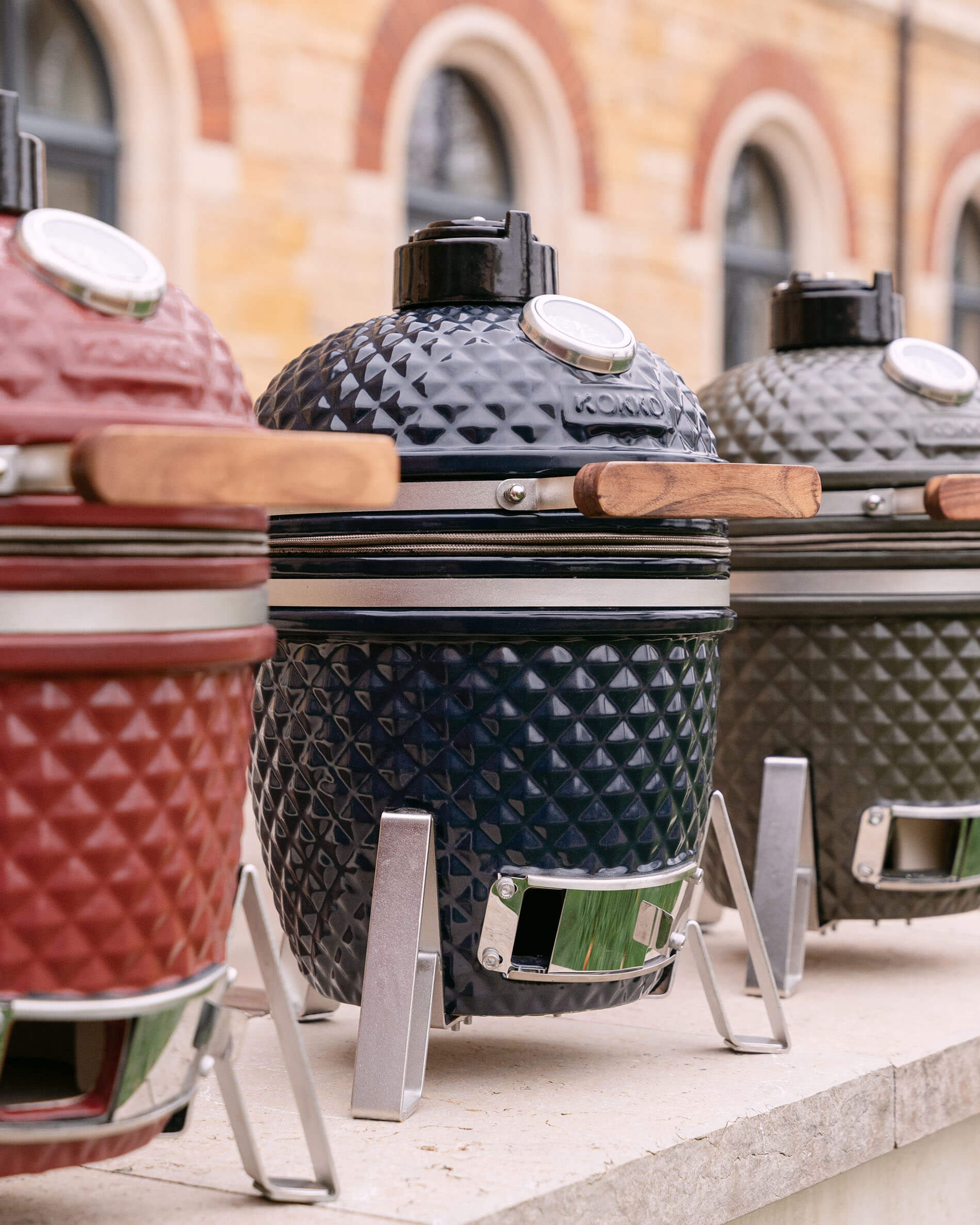 Kamados Kokko alignés en céramique texturée, présentés en plusieurs coloris avec poignées en bois et supports métalliques.