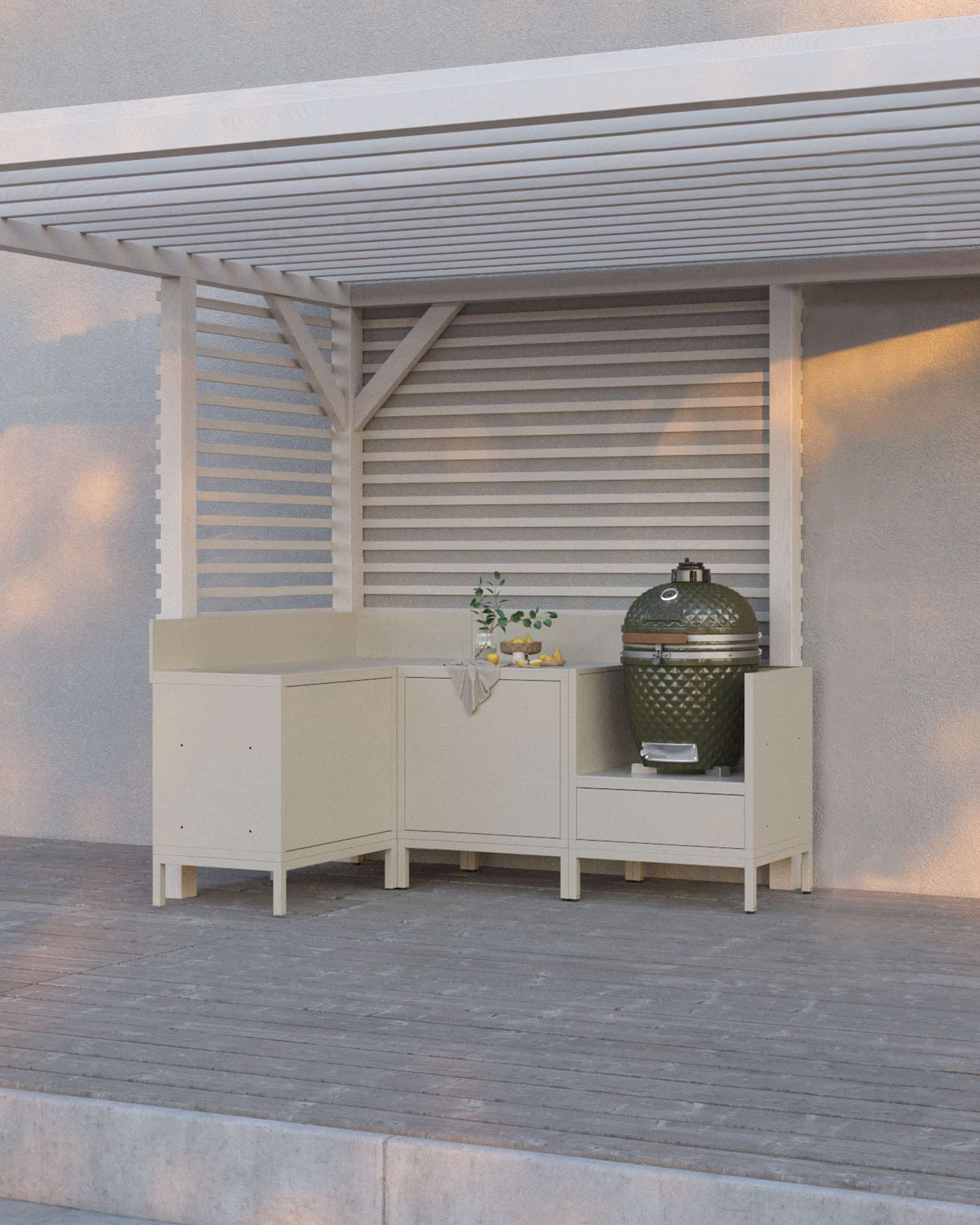 Cuisine pilotis coloris crème composée de quatre modules avec angle, plan de travail continu et emplacement dédié au kamado, installée sous une pergola blanche sur terrasse en bois.