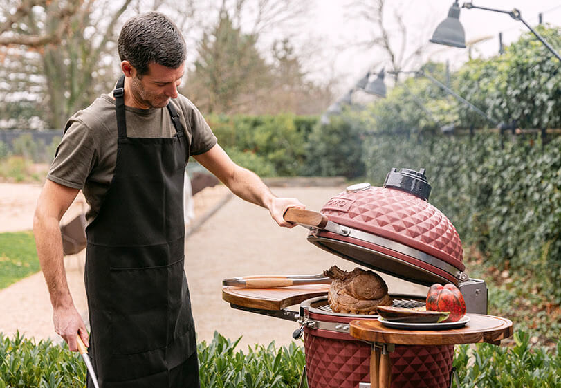 Roast cooking in a red Kokko kamado, with the lid open and food being handled outdoors.